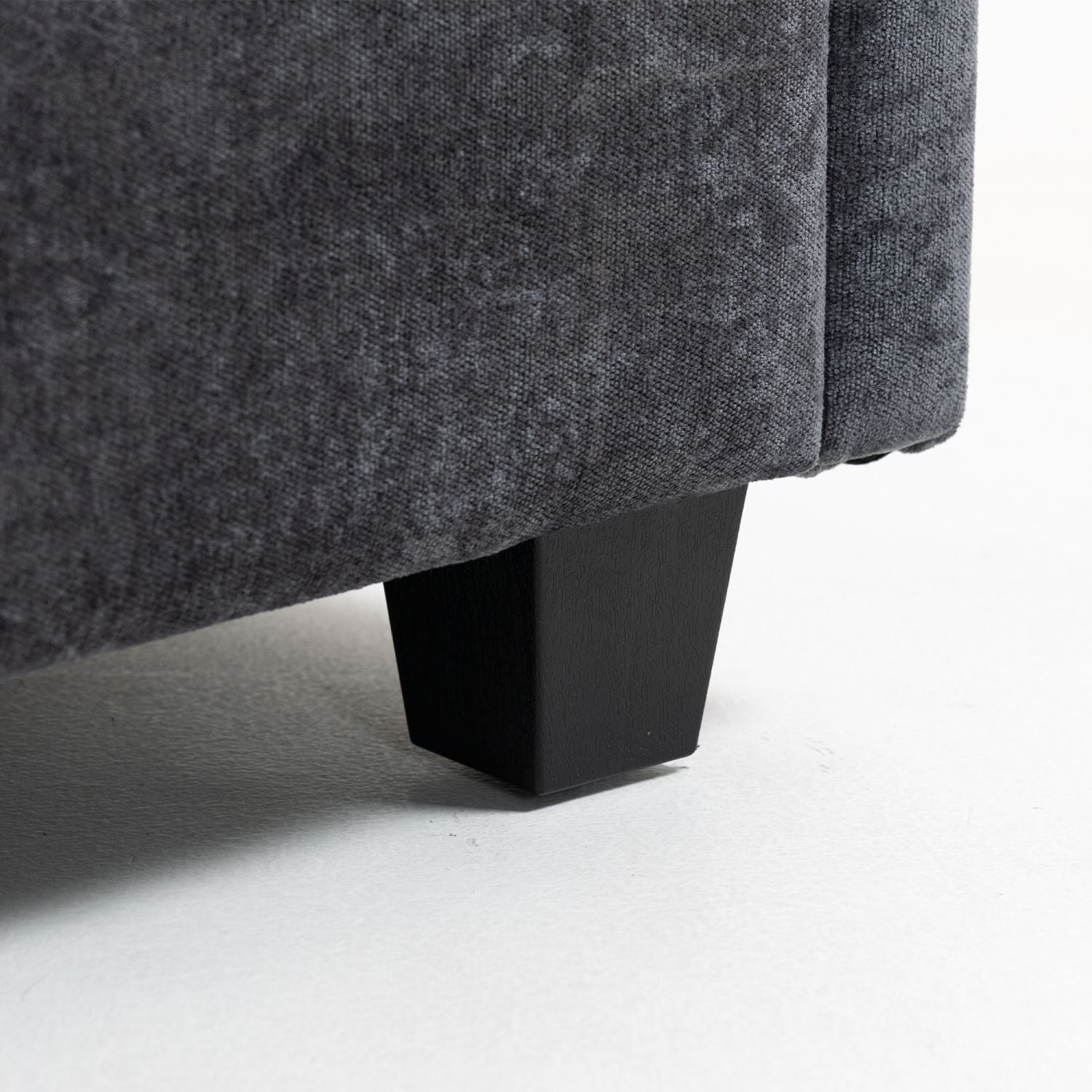 Close-up of dark grey chenille fabric sofa leg with black tapered wooden base on white floor, part of ultra-soft XXXL 3-seater sofa